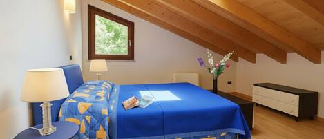 A bedroom with a sloped ceiling, a bed with a blue bedspread, a bedside table with a lamp, a nightstand, and a dresser.