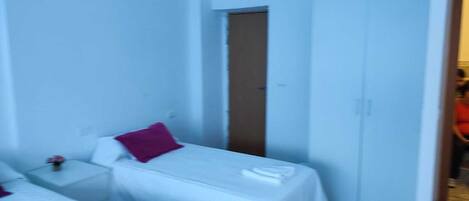 A small, single-room hotel room with two beds, a door, and a nightstand with a lamp.
