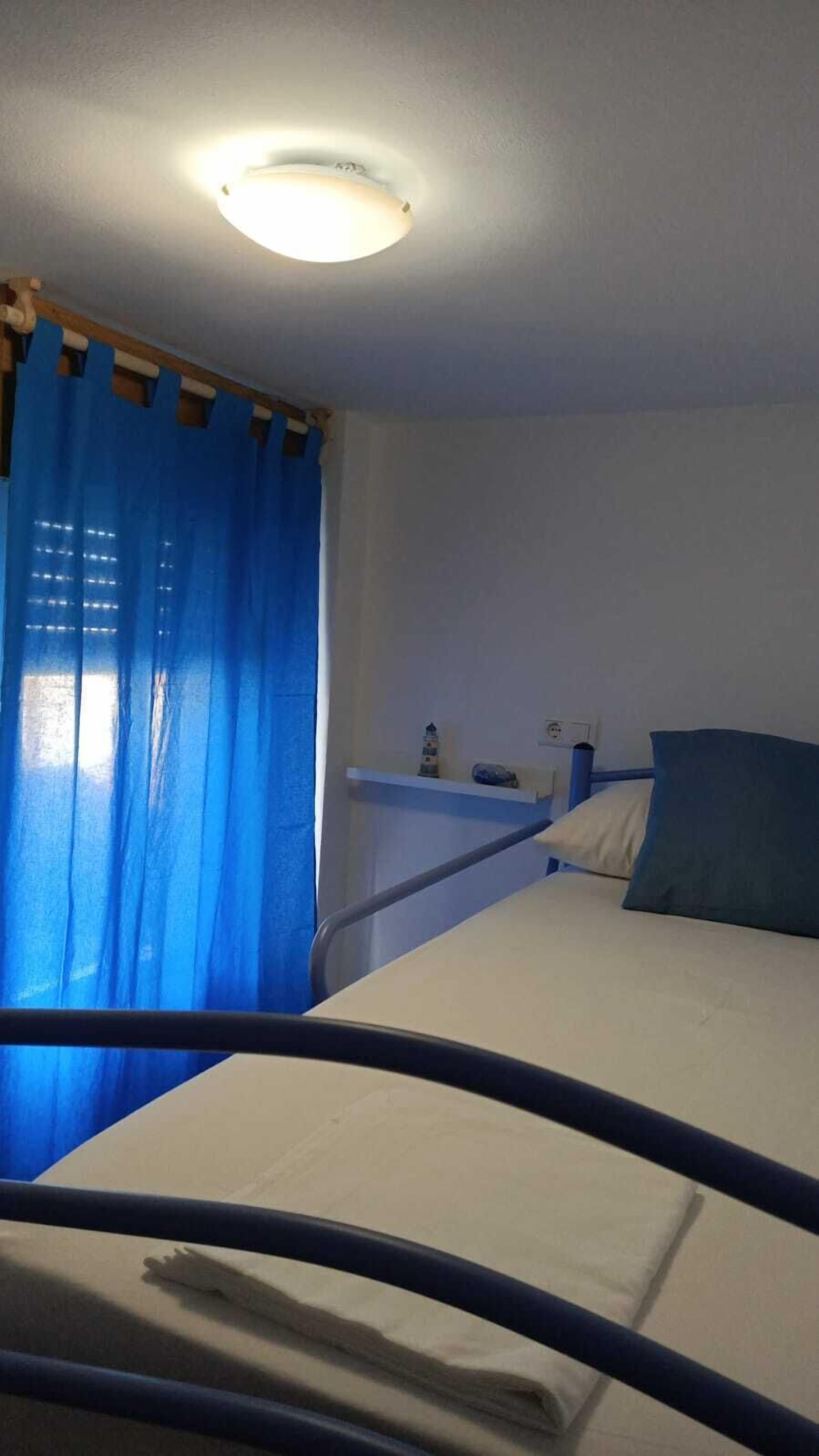 Una habitación con cama, mesita de noche, lámpara y ventana con cortinas azules.