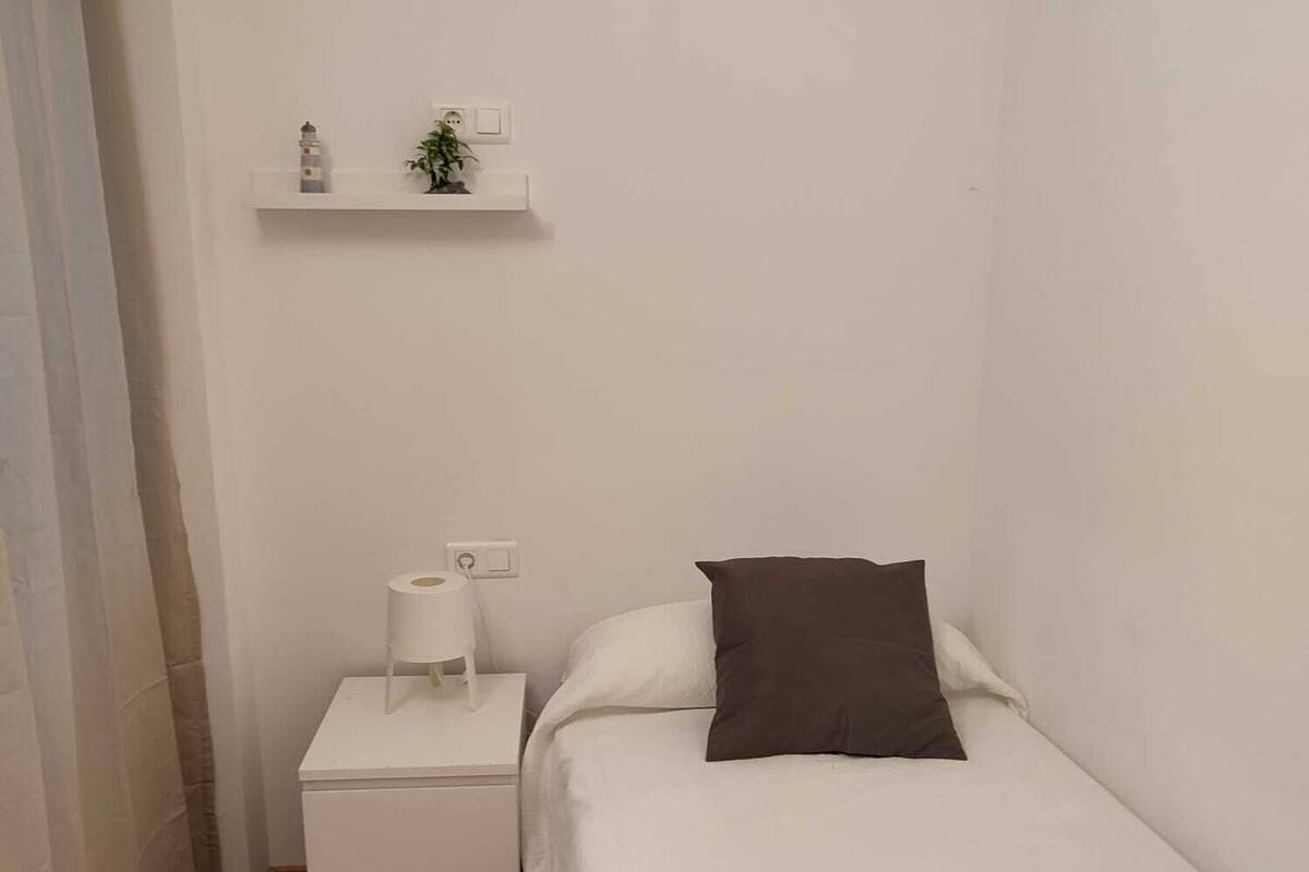 A small, clean room with a single bed, a nightstand with a lamp, a shelf with a plant, and a wooden floor.
