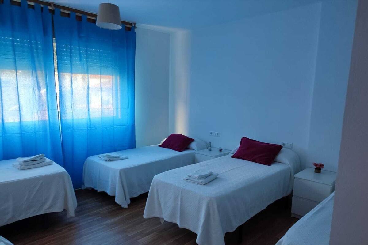 A hotel room with two single beds, a small bedside table with a lamp, and a window with blue curtains.