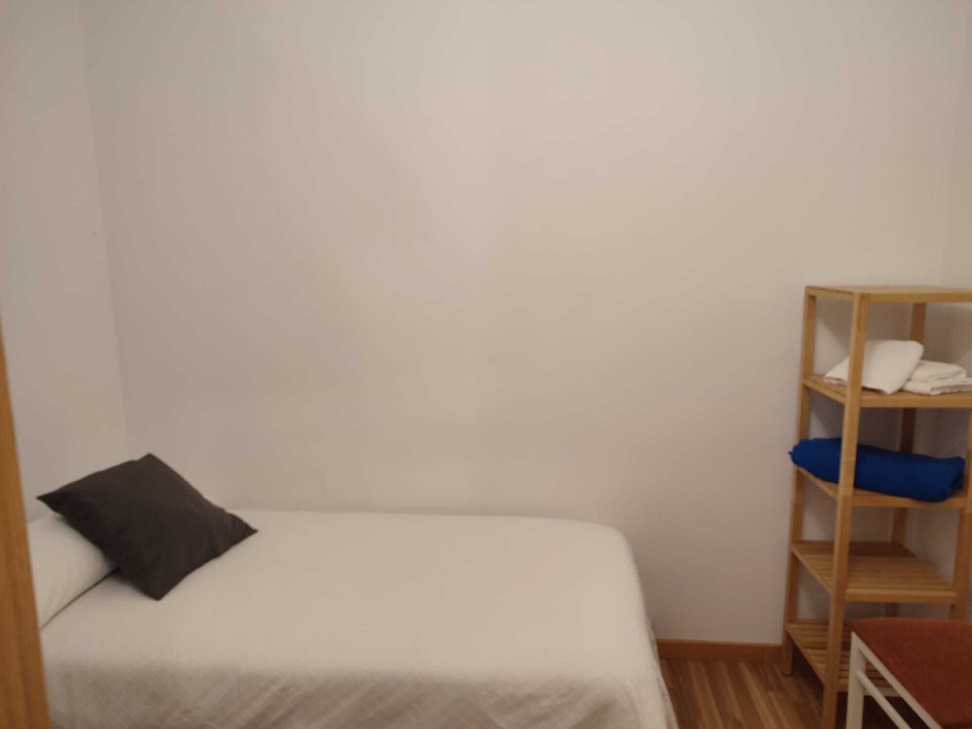 Una habitación pequeña y sencilla con una cama individual, un estante de madera con ropa doblada y una mesita.