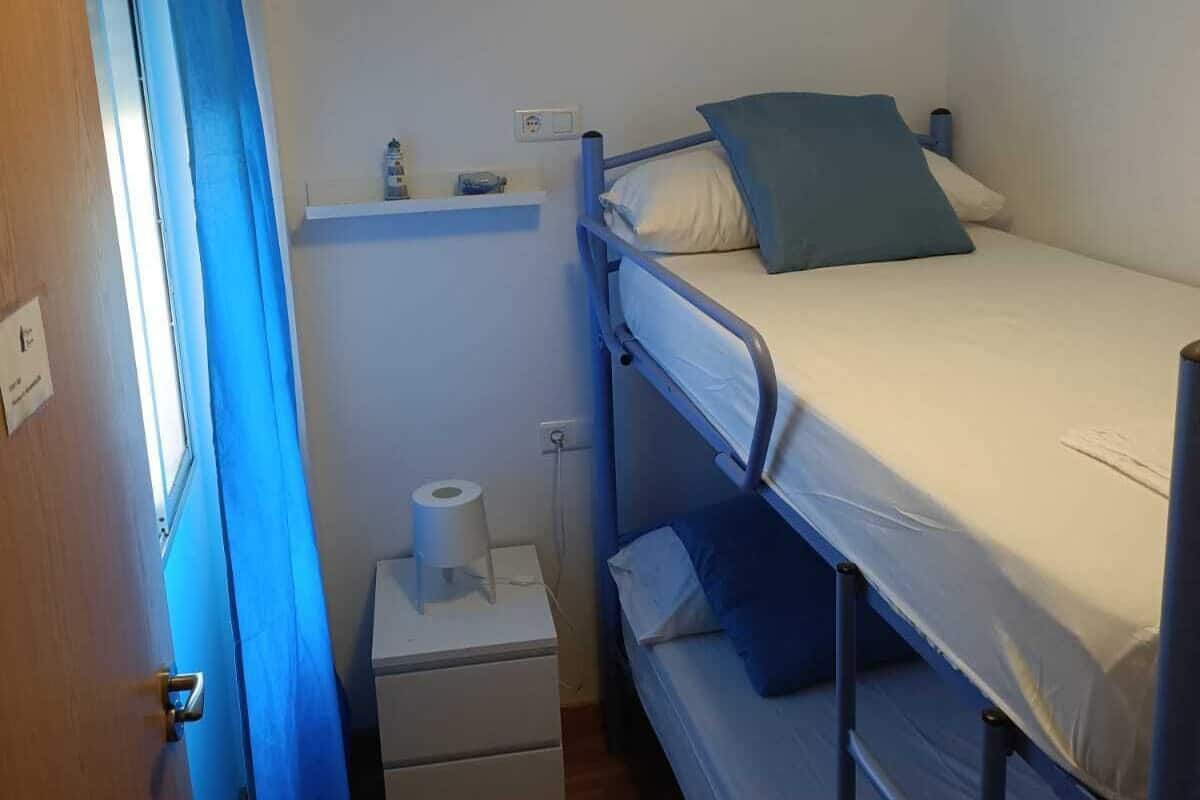 A small, compact room with bunk beds, a small shelf with toiletries, a toilet, and a window with blue curtains.