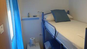 Una habitación pequeña y funcional con literas, un estante con artículos de aseo, un inodoro y una ventana con cortinas azules.