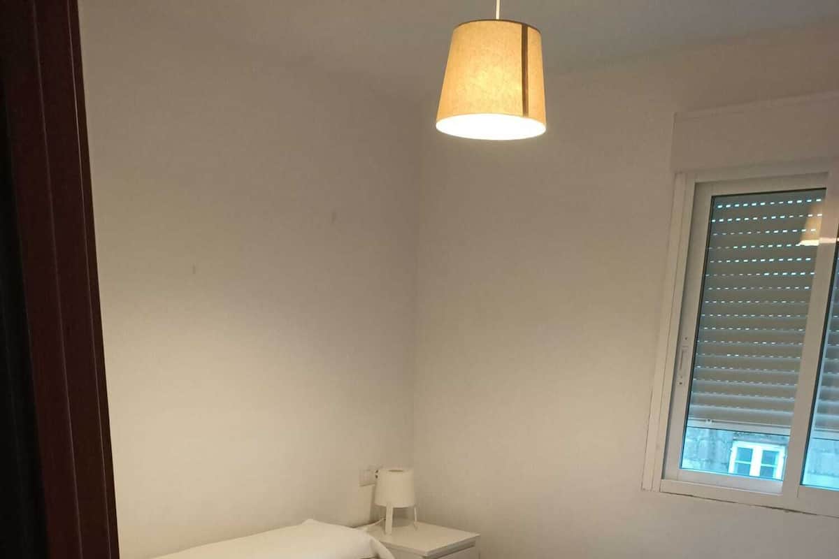 A single bed with a white bedspread, a bedside lamp, and a window with blinds.