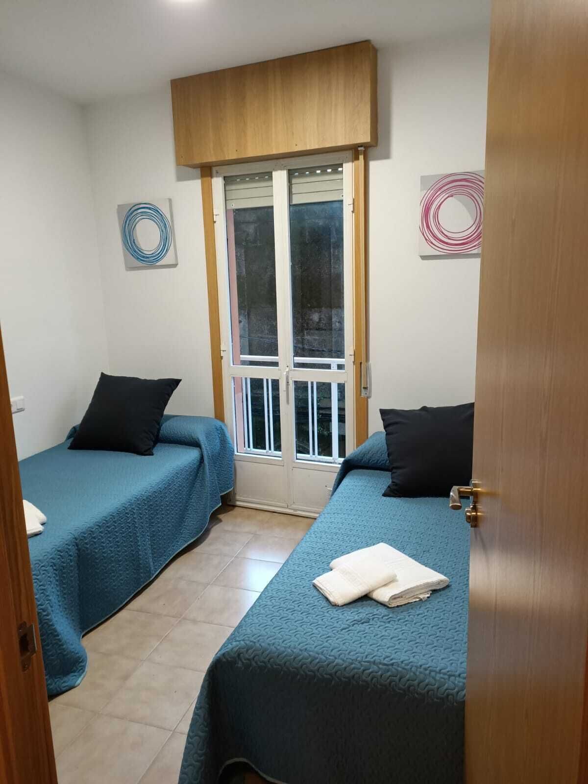 Una habitación con dos camas individuales, cada una con una colcha azul y almohadas negras. Hay una puerta de madera con un panel de vidrio y una obra de arte circular en la pared.