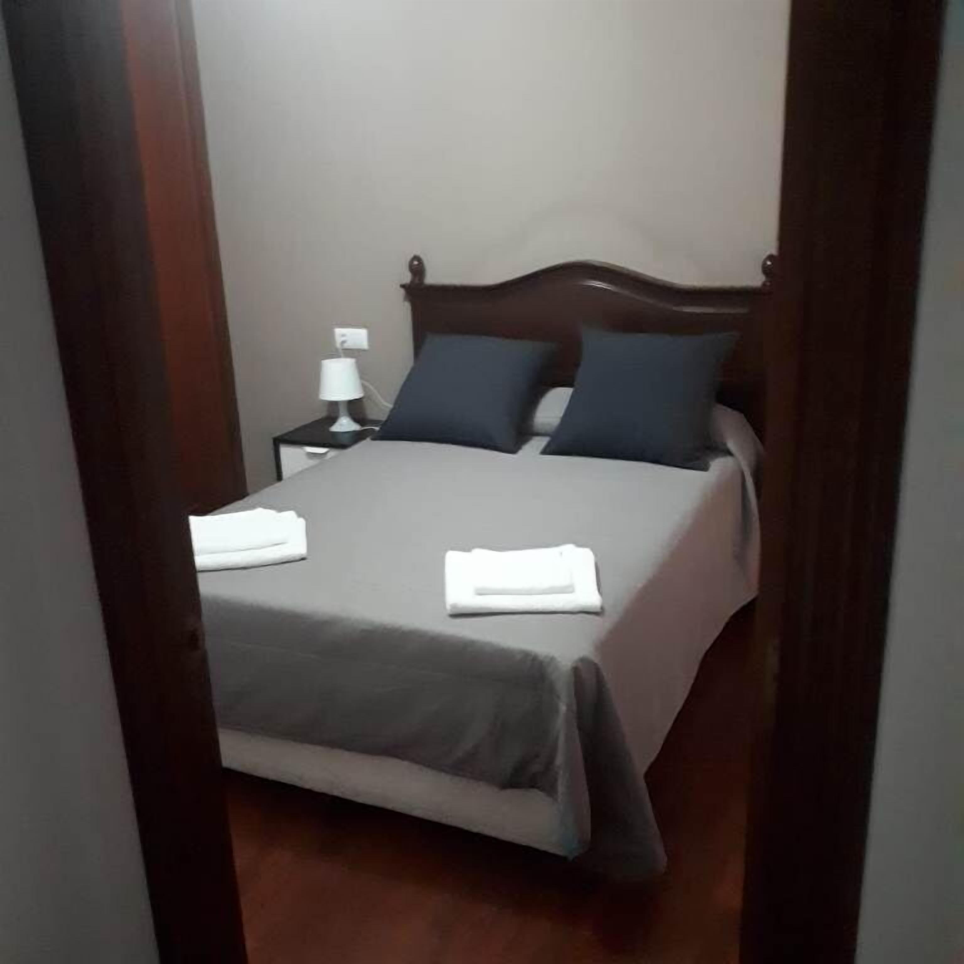 Una cama bien hecha con una colcha gris y dos almohadas, una mesita de noche con una lámpara y un cabecero de madera.
