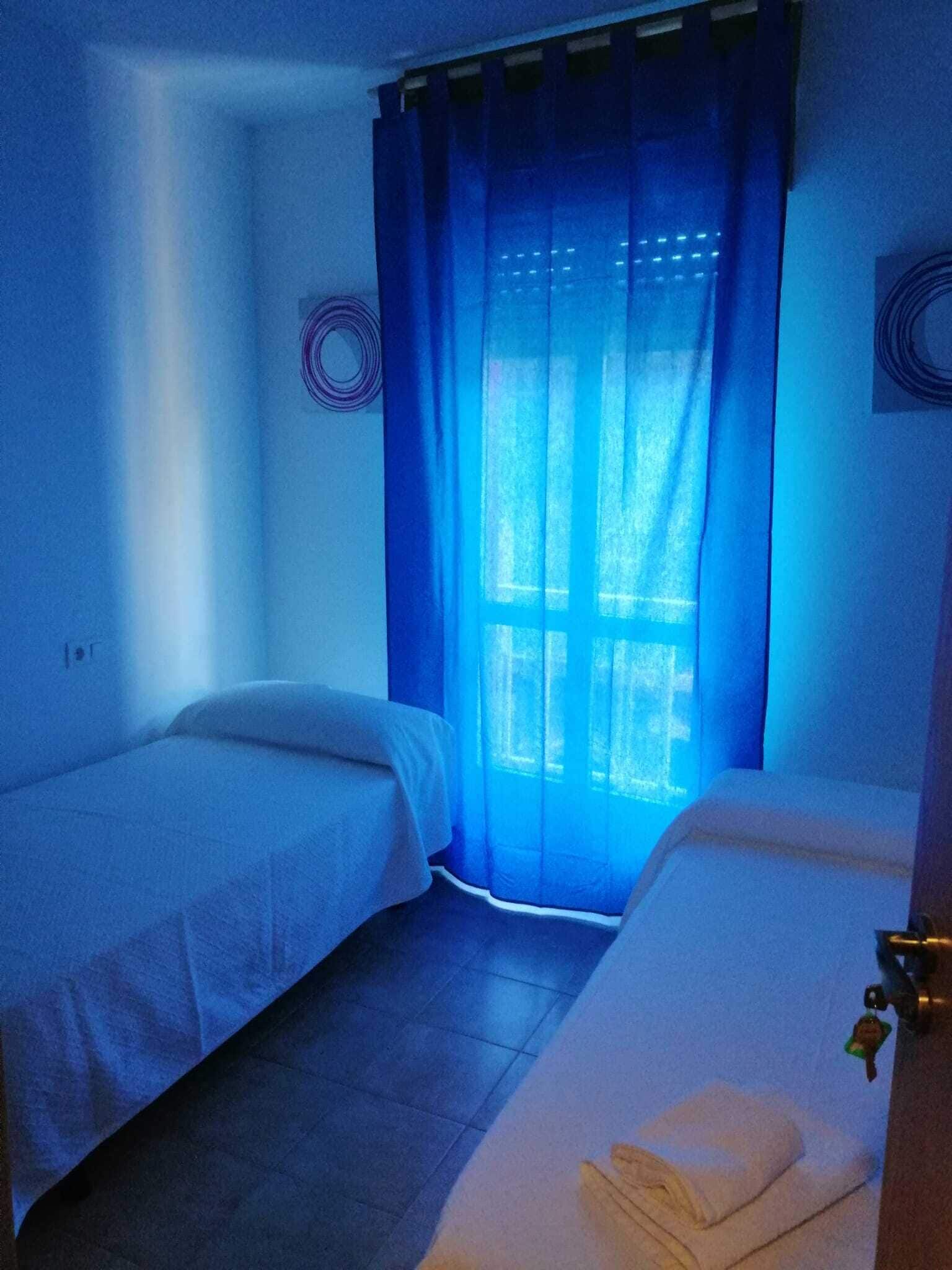 Una habitación pequeña y tenuemente iluminada, con dos camas individuales, una ventana con cortinas azules y una puerta con un llavero.