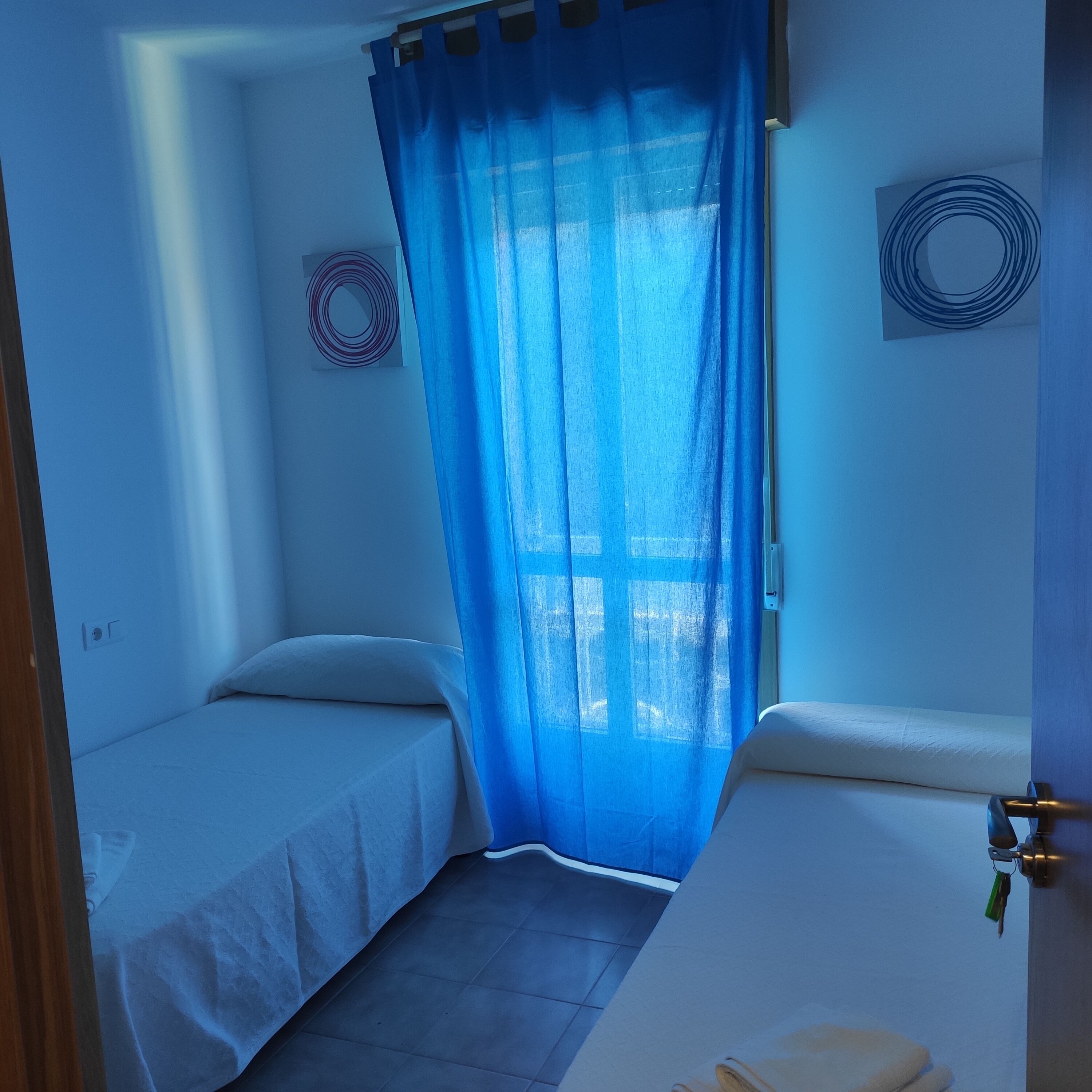 Habitación pequeña de hotel, con cama, una mesita y ventana con cortinas azules.