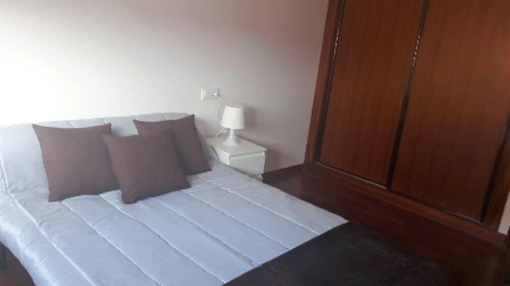 Una cama con ropa de cama blanca y almohadas marrones, una mesita de noche con lámpara, y un armario de madera.