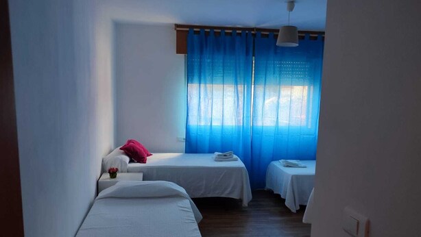 A room with two single beds, a small table with a red pillow, and a window with blue curtains.