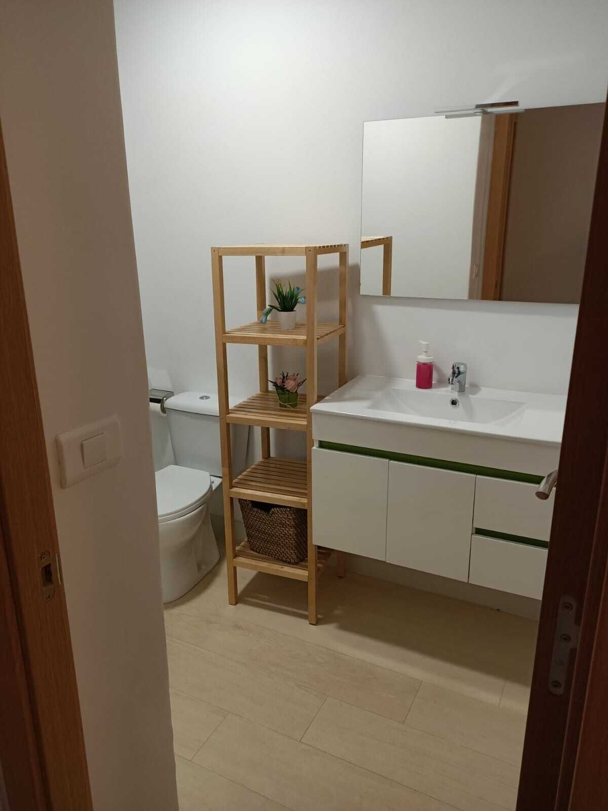 Un baño con un estante de madera, un inodoro, un lavamanos con espejo y una franja verde en la encimera.