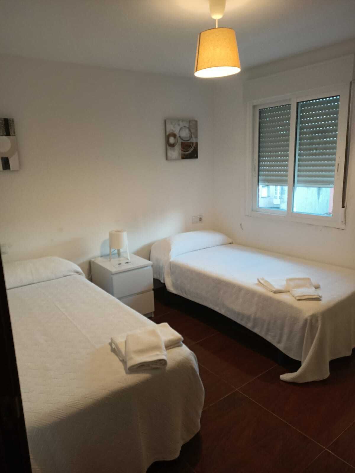 A room with two single beds, a nightstand with a lamp, a window with blinds, and a ceiling light.