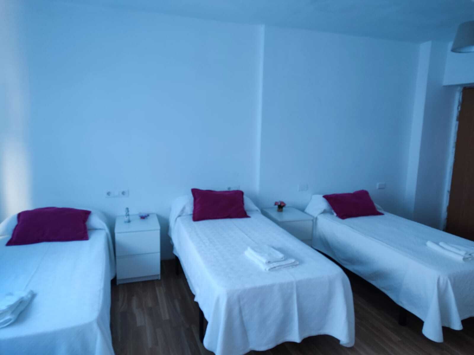 Habitación de hotel con tres camas individuales, cada una con una colcha blanca y almohada roja. Hay mesitas de noche blancas con lámparas y una pequeña mesa con un jarrón de flores.