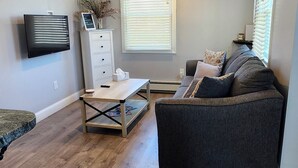 Una sala de estar con un sofá gris oscuro, una mesa de centro, un televisor montado en la pared y una ventana con persianas.