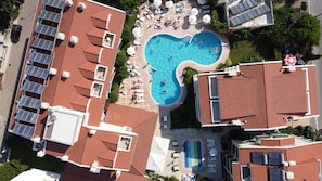 An aerial view of a hotel with a central swimming pool surrounded by sun loungers and umbrellas, solar panels on the rooftops, and a mix of red-tiled roofs and white walls.