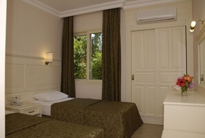 A hotel room with two beds, a bedside table with a phone, a window with curtains, a dresser with a vase of flowers, and an air conditioning unit.