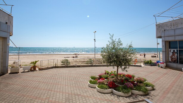 A paved walkway with a view of the beach and ocean, a small tree, and a building with a sign. - Hotel Kojku (Durrës)