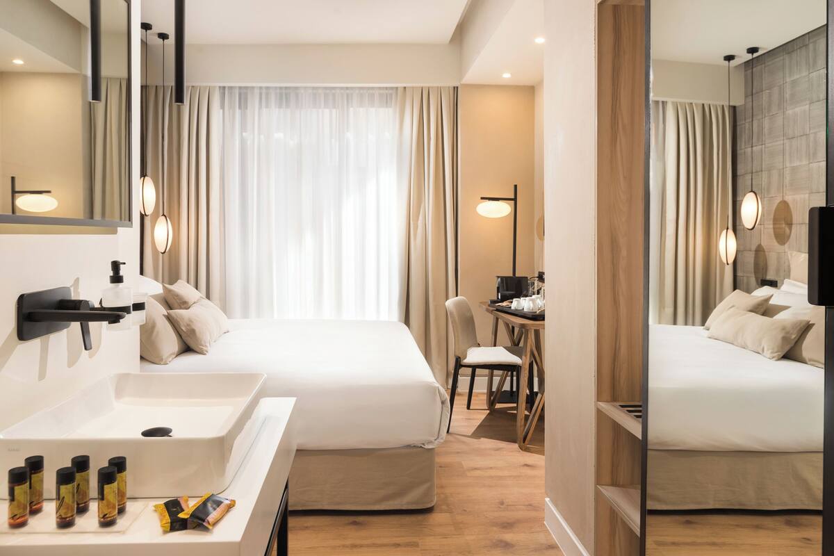 A hotel room with a large bed, a desk, a chair, a bathroom with a sink and mirror, and a large window with curtains.
