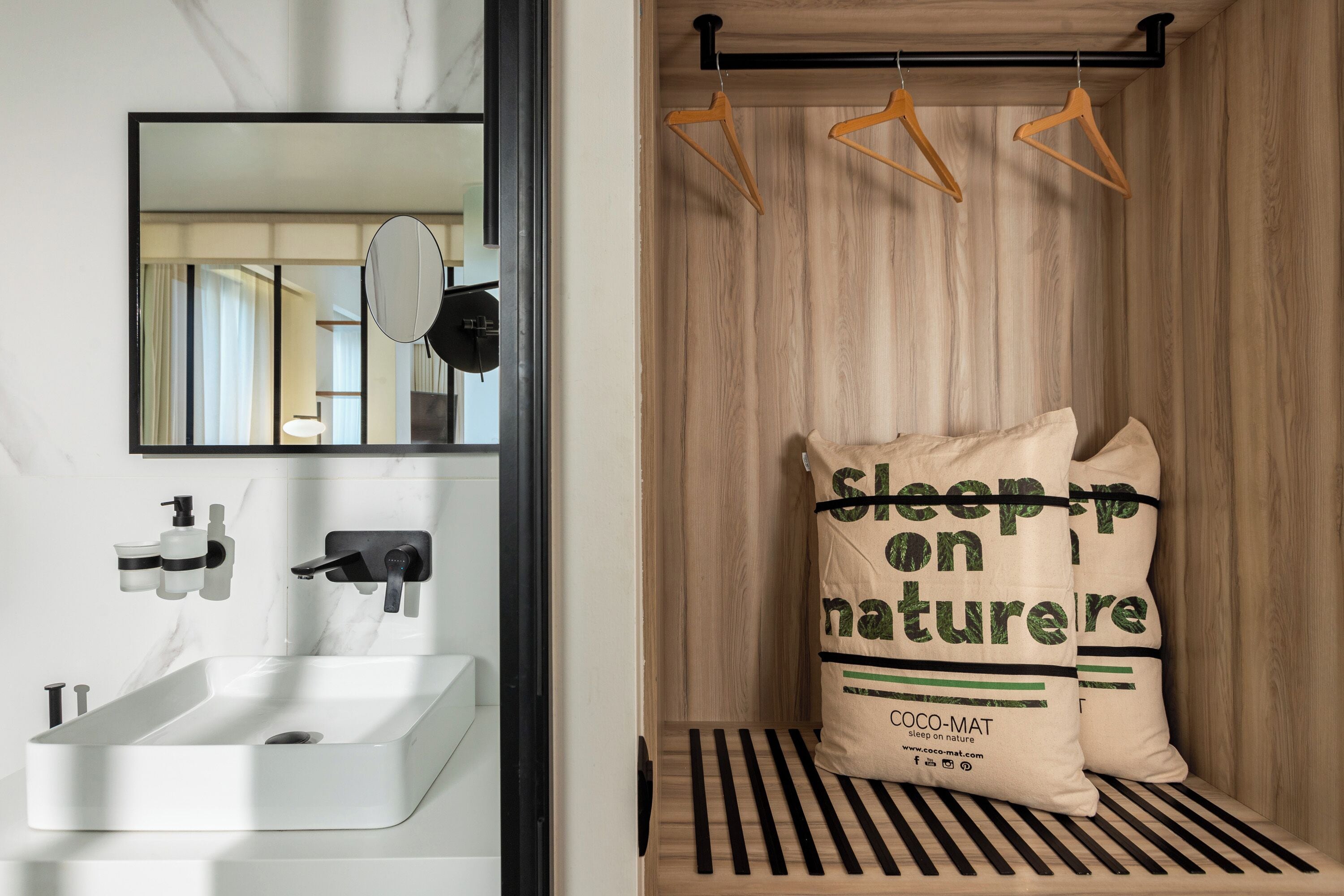 A modern bathroom with a white sink, mirror, and wooden paneling. There are two hanging clothes racks and a bed with a striped mattress and pillows with "Sleep on Nature" branding.