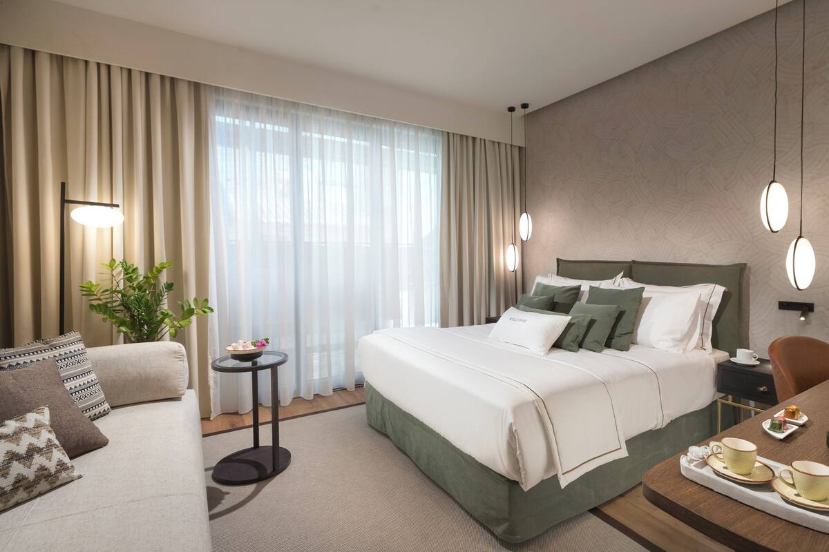 A hotel room with a large bed, a bedside table with a coffee maker, a small table with cups, and a potted plant.