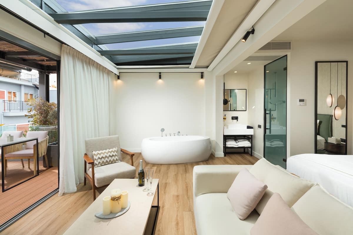 A modern hotel room with a large skylight, a bathtub, a sofa, and a dining area with a table and chairs.
