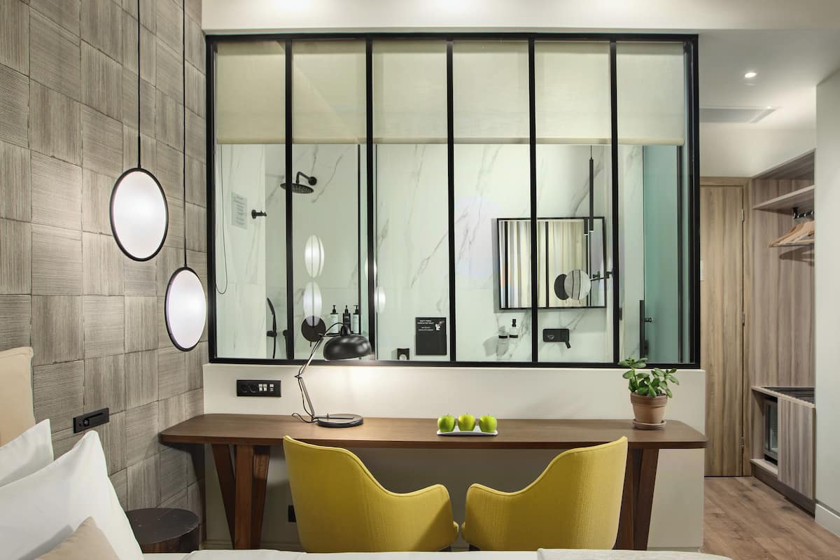A modern hotel room with a large window, a wooden desk, a bed, and a mirror.