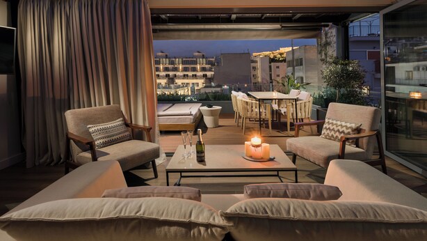 A modern living room with a sofa, chairs, a table with candles, and a view of the city at dusk. - The Residence - Christokopidou Hotel & SPA (Athens)