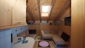 A cozy attic room with wooden walls and a sloped ceiling. There is a sofa, a small table with a doily, and a window providing natural light. - Chalet Häsi 3 (4) room attic apartment (Hasliberg-Wasserwendi)