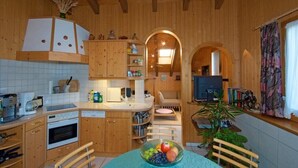 A cozy kitchen with wooden cabinets, a dining table with a fruit bowl, and a television. - Chalet Häsi 3 (4) room attic apartment (Hasliberg-Wasserwendi)