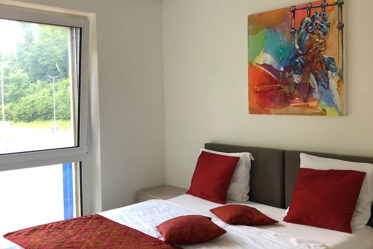 Una moderna camera d'albergo con un letto grande, biancheria rossa e bianca, un quadro astratto colorato sulla parete e una finestra con vista sul verde.