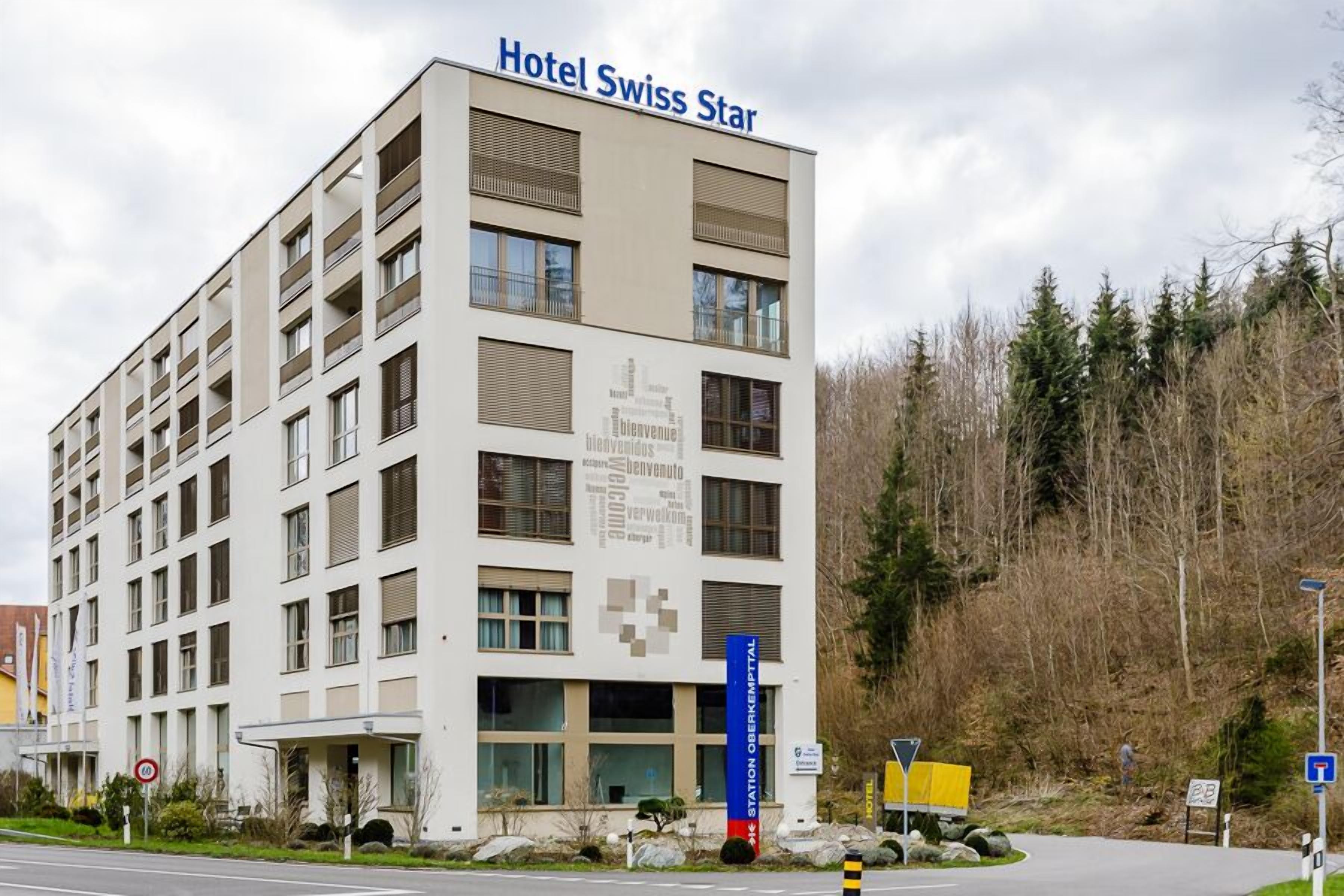 A multi-story hotel named Hotel Swiss Star with a modern design, featuring a prominent entrance and a sign indicating a station or transportation hub nearby.