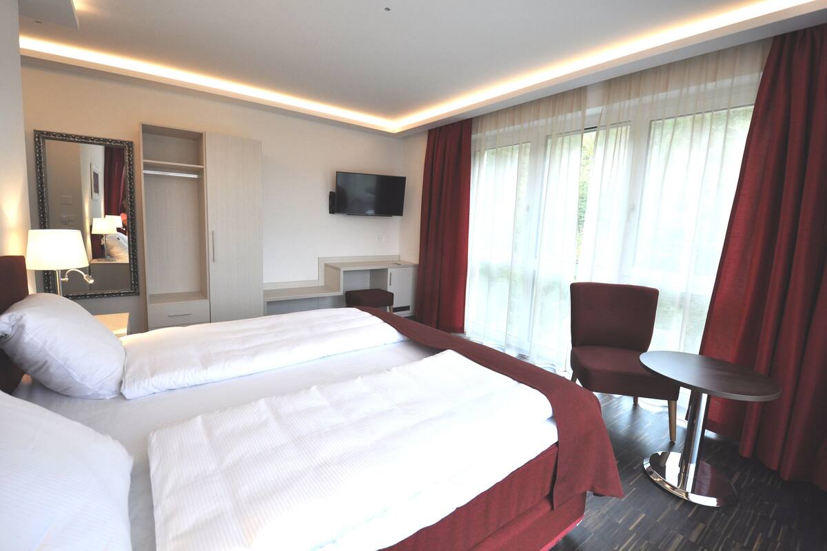 Camera d'albergo con due letti, una scrivania, una sedia, una televisione e ampie finestre con tende.