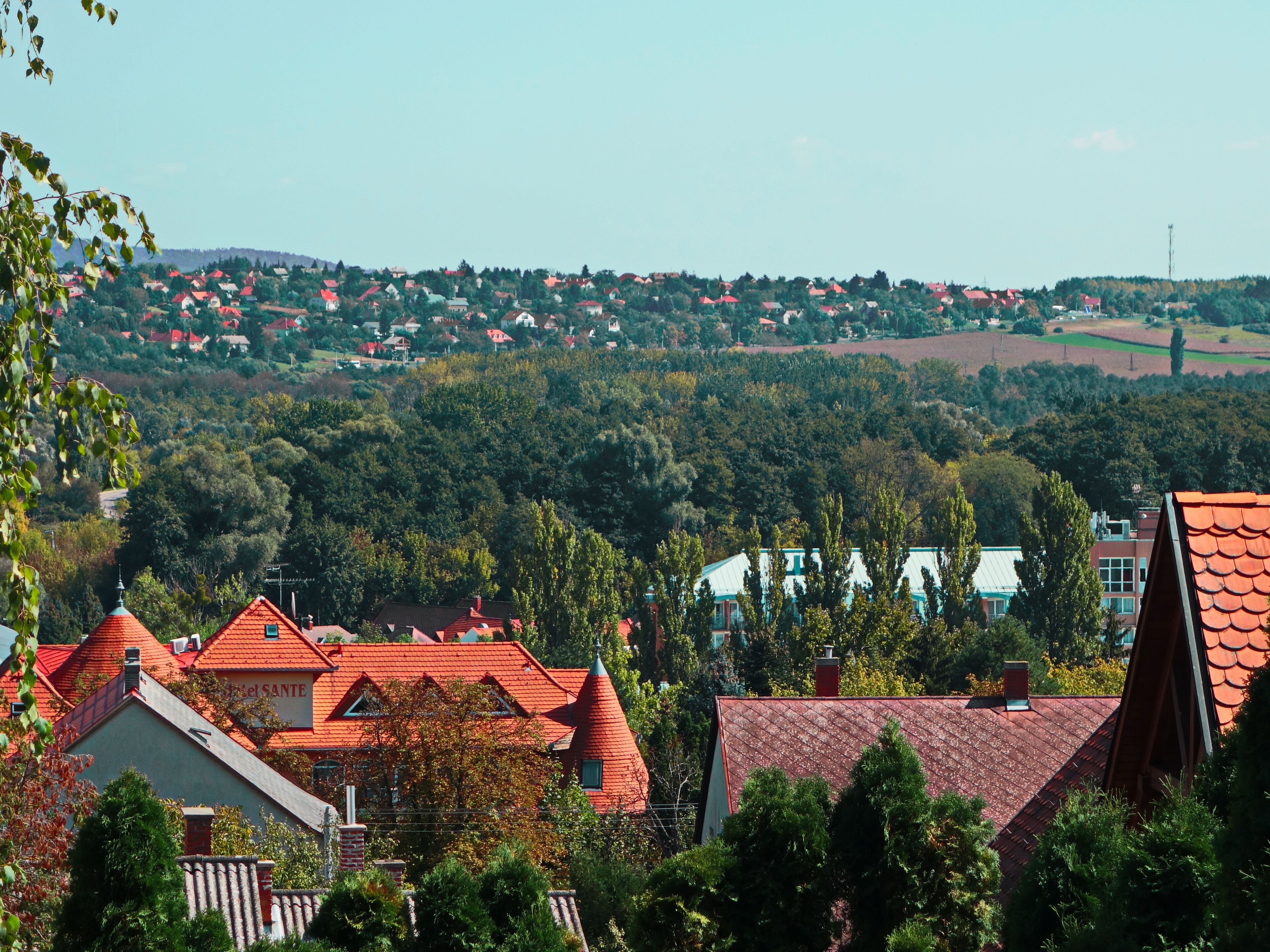 A residential area with houses having red roofs, surrounded by greenery and hills in the background.