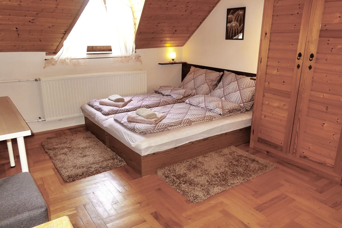 A bedroom with a wooden ceiling, a bed with patterned bedding, a nightstand, a wooden wardrobe, and a radiator.