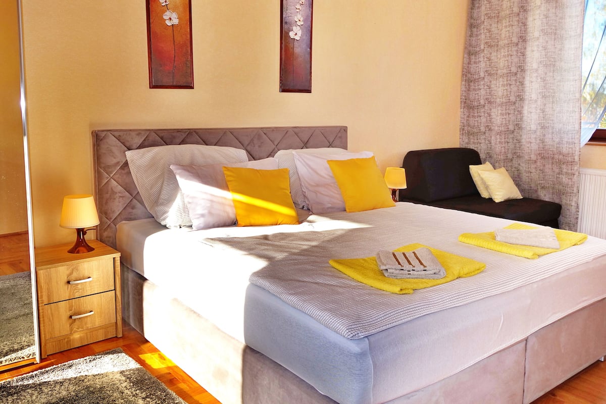 A neatly made bed with pillows, a bedside table with a lamp, a chair, and artwork on the wall.