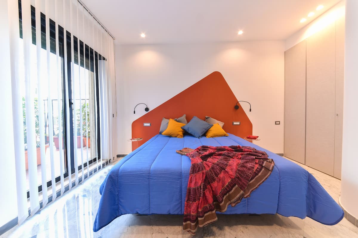 A modern bedroom with a large bed, a red accent wall, and a view through large windows.