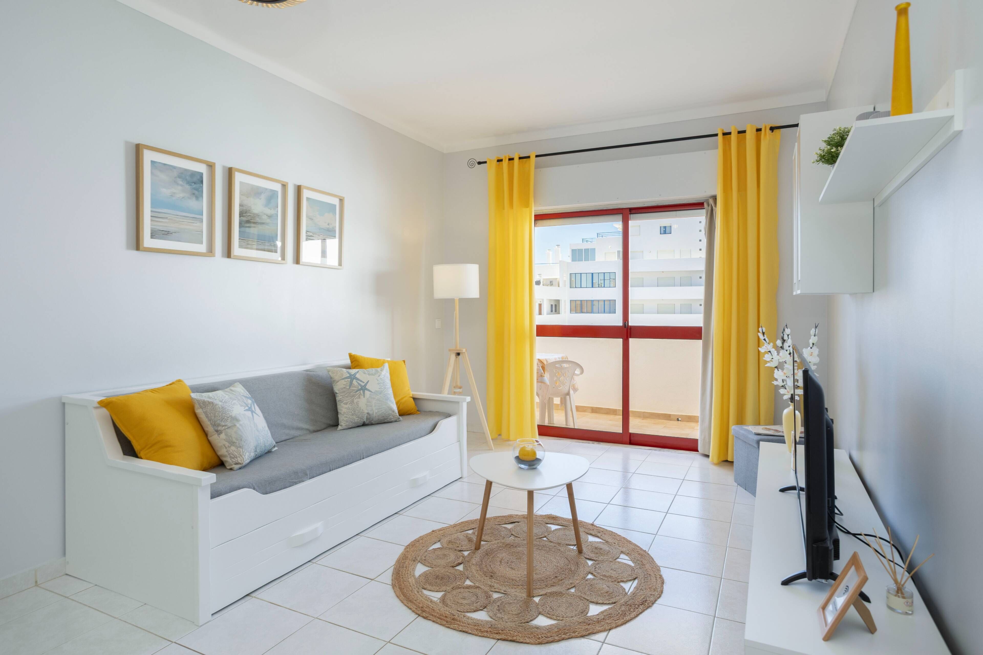 A modern living room with a white sofa, yellow cushions, a small round table, and a TV on a white stand.