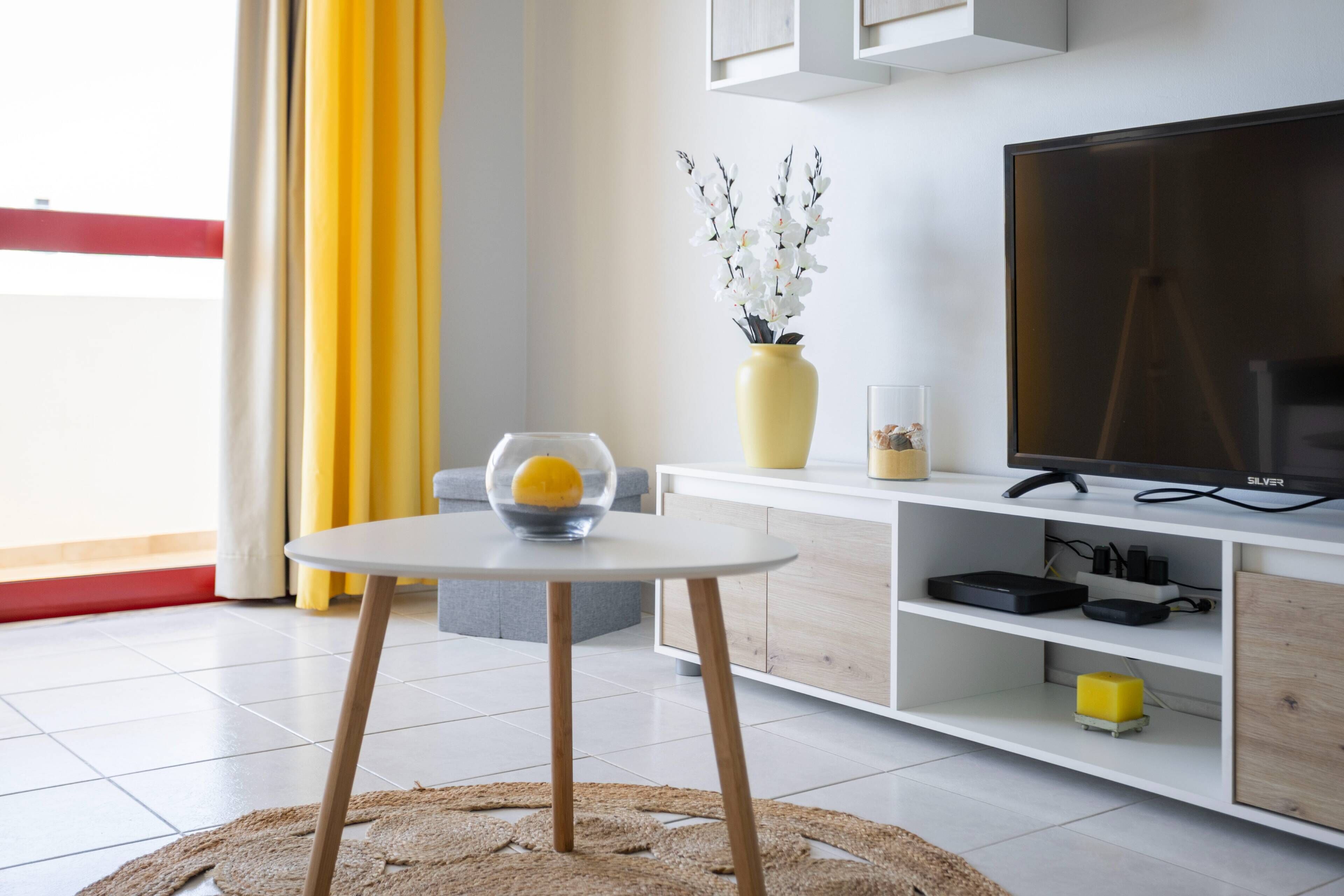 Un salón moderno con un mueble blanco para el televisor, una mesa de vidrio con un limón y un jarrón de flores blancas.