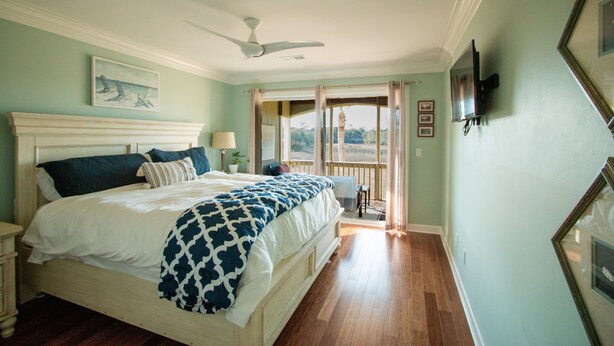 A bedroom with a large bed, a ceiling fan, a nightstand, a lamp, and a view of the outdoors through a door. - Gated Secluded Marsh View (Johns Island)