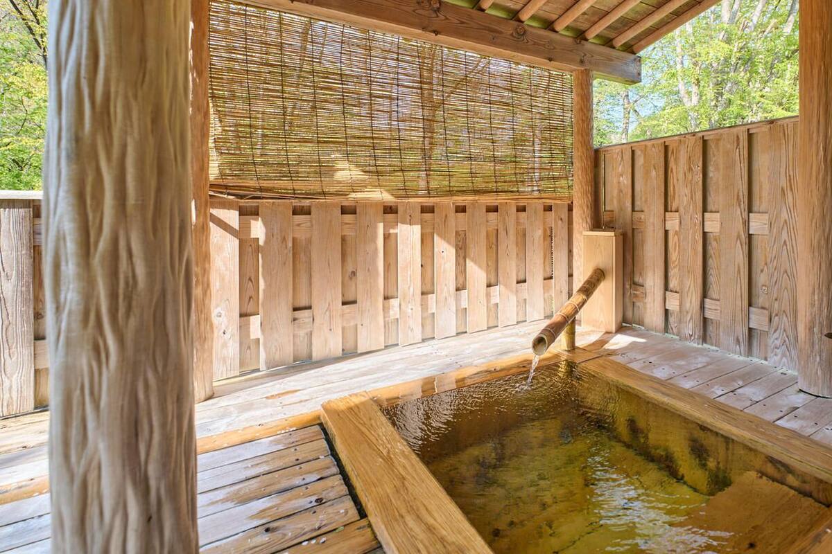 Un jacuzzi exterior de madera con separador de bambú, terraza de madera y un pequeño taburete de madera.