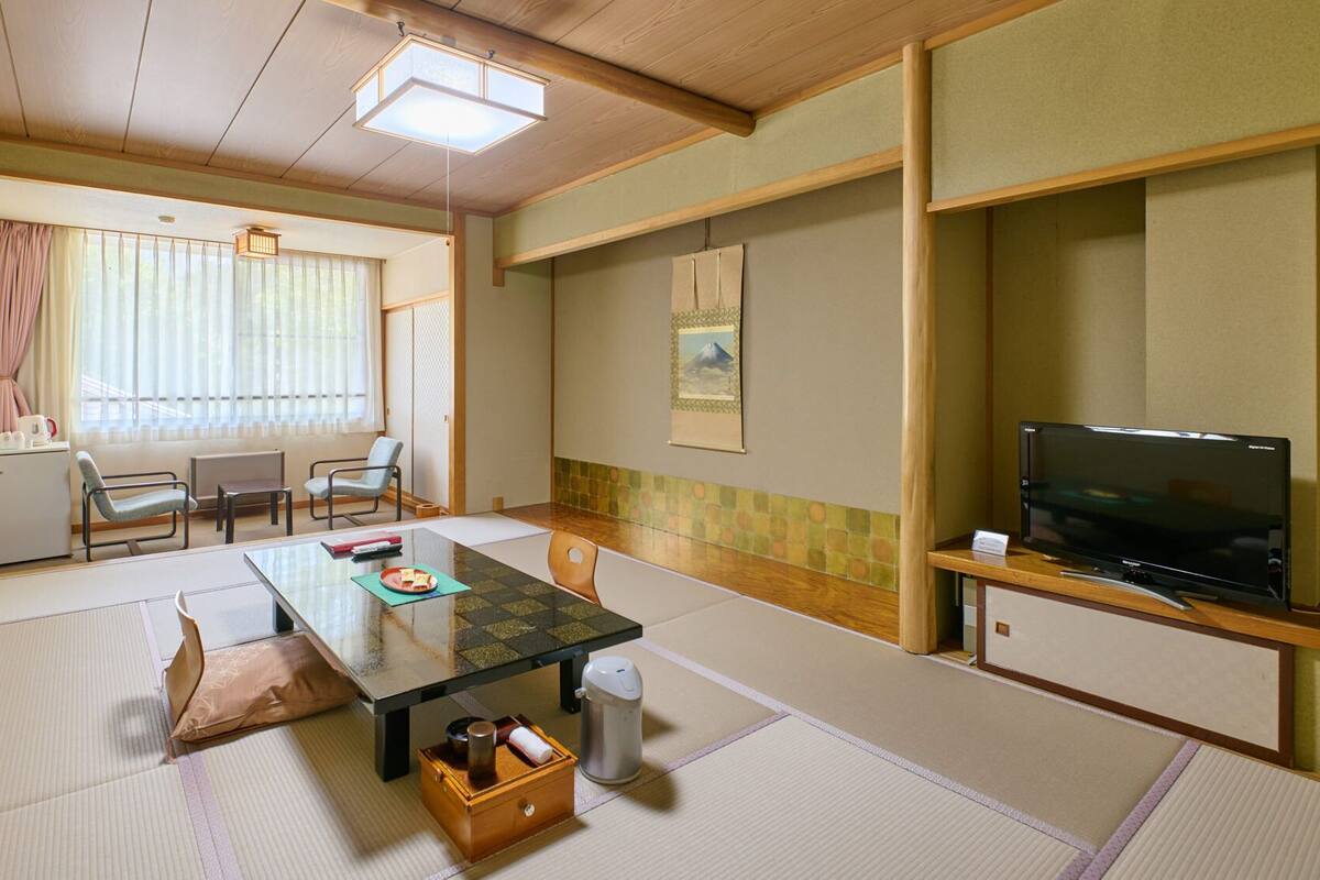Una habitación tradicional japonesa con suelo de tatami, una mesa baja, sillas, un televisor y un refrigerador pequeño.