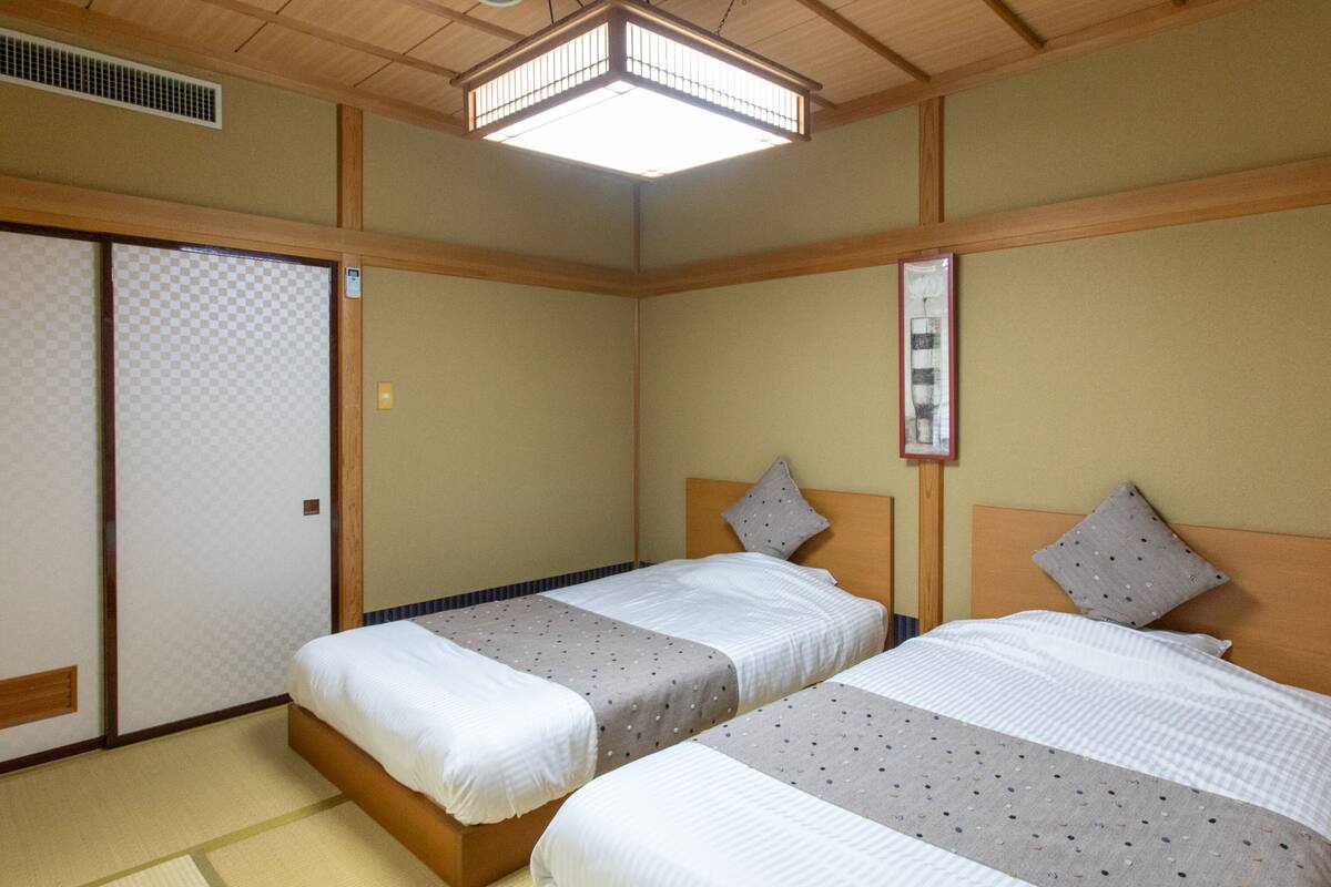 Habitación de hotel con dos camas, esteras de tatami, un biomblo shoji y un ventilador de techo.