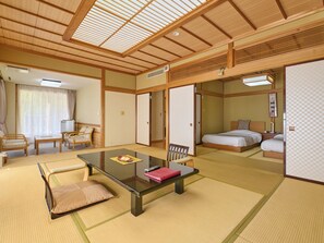 Una habitación tradicional japonesa con suelo de tatami, una mesa baja con cojines, una cama con cabecero de madera y puertas correderas.