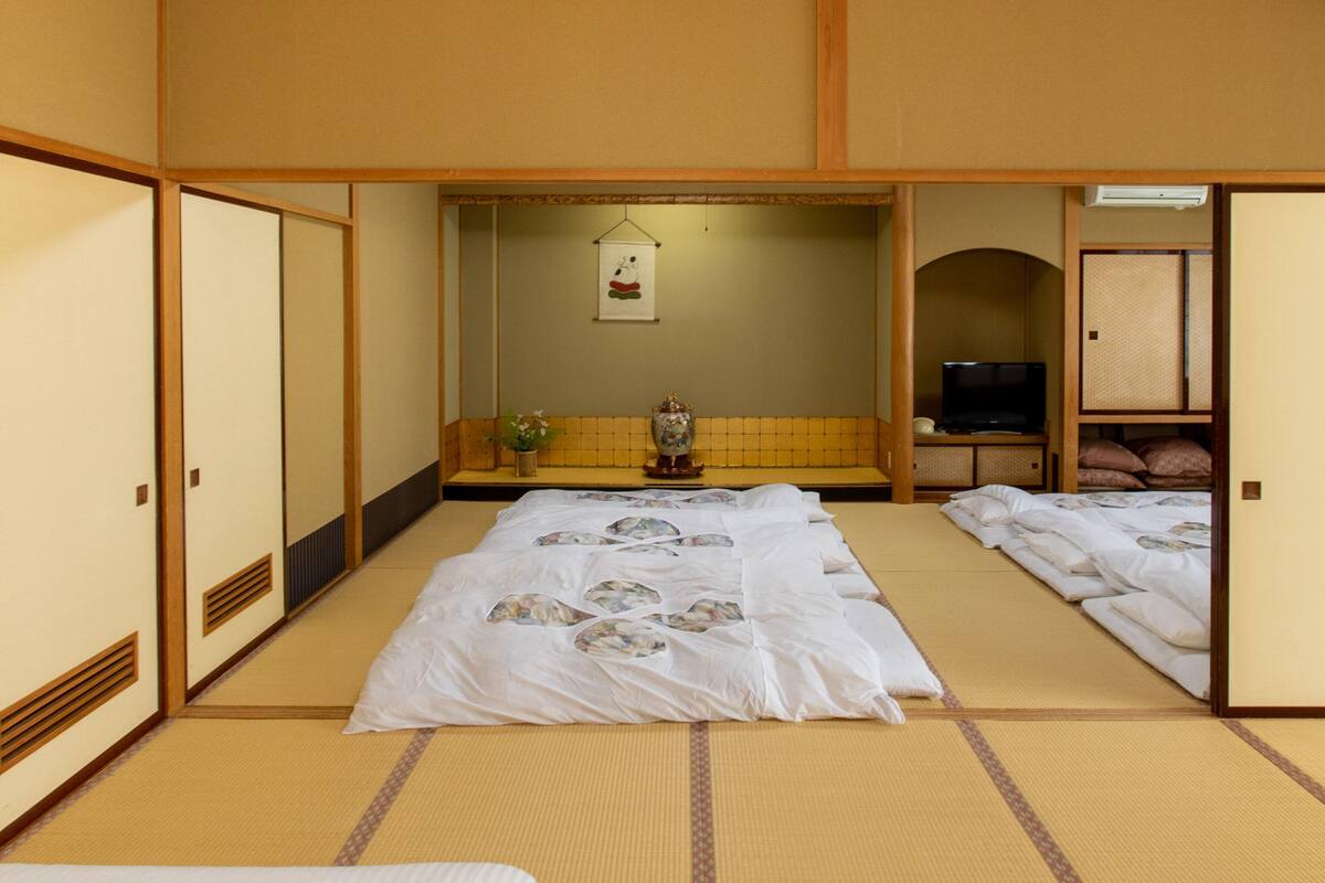 Una habitación japonesa tradicional con suelo de tatami, una cama con colcha estampada, un televisor y un pequeño santuario con un adorno colgante.