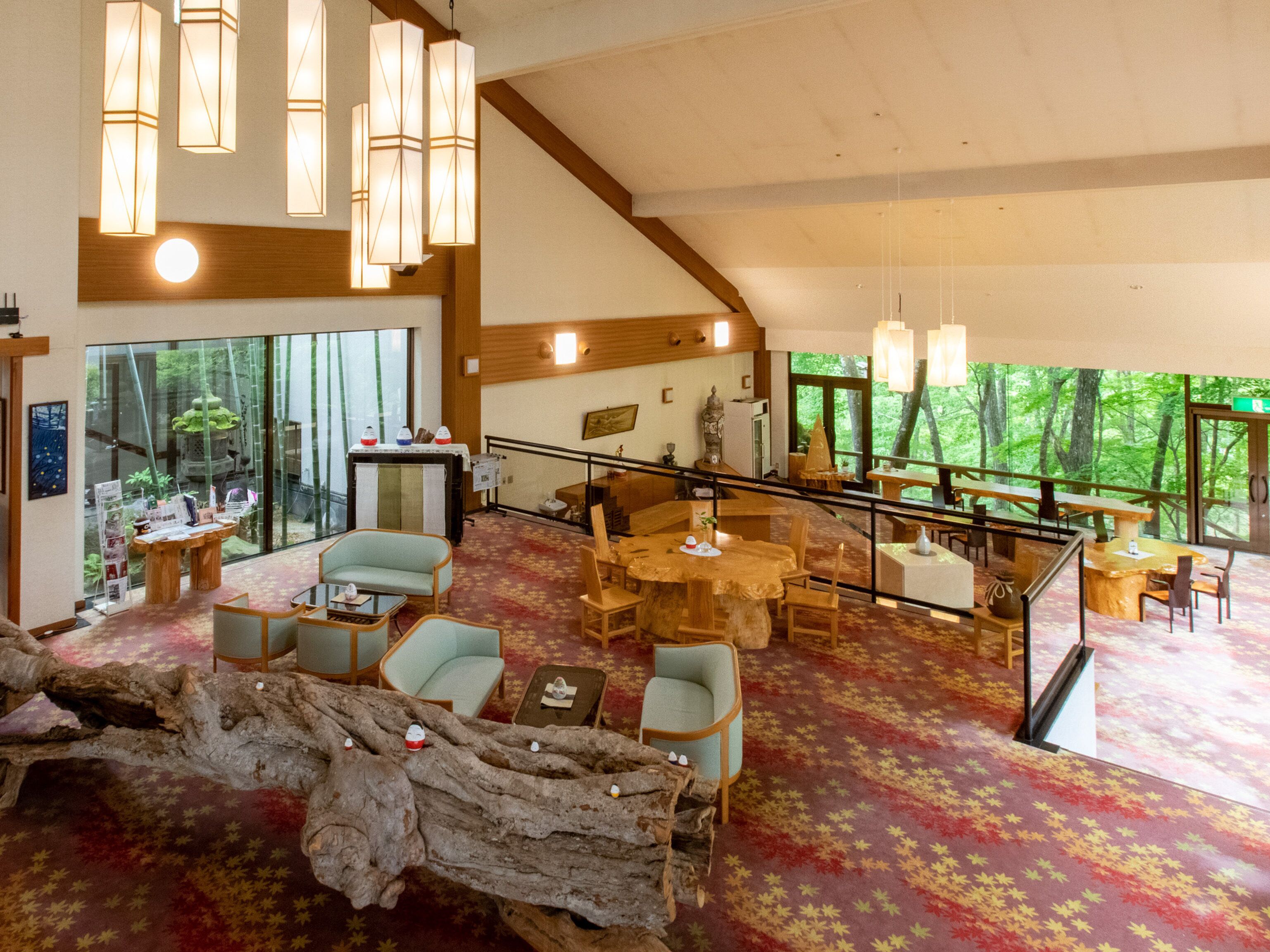 A spacious room with a high ceiling, wooden beams, and large windows offering a view of the forest. There are multiple seating areas with tables and chairs, and a large tree trunk decoration in the center.
