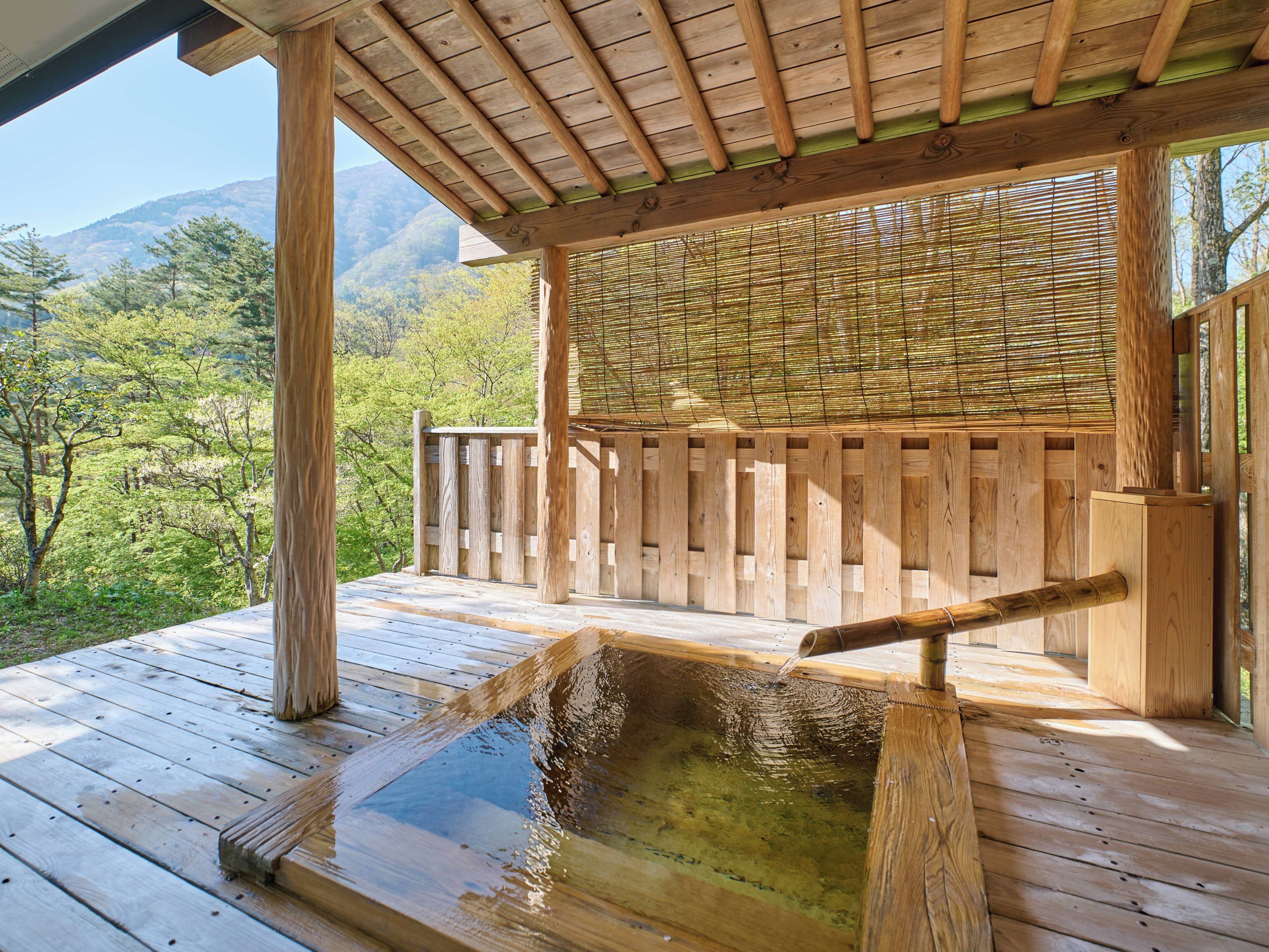 A wooden deck with a hot spring, surrounded by a wooden fence and bamboo blinds, overlooking a forested mountain area.