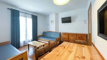 Una habitación compacta con una mesa de comedor de madera, un sofá azul, un televisor montado en la pared y una ventana con cortinas.