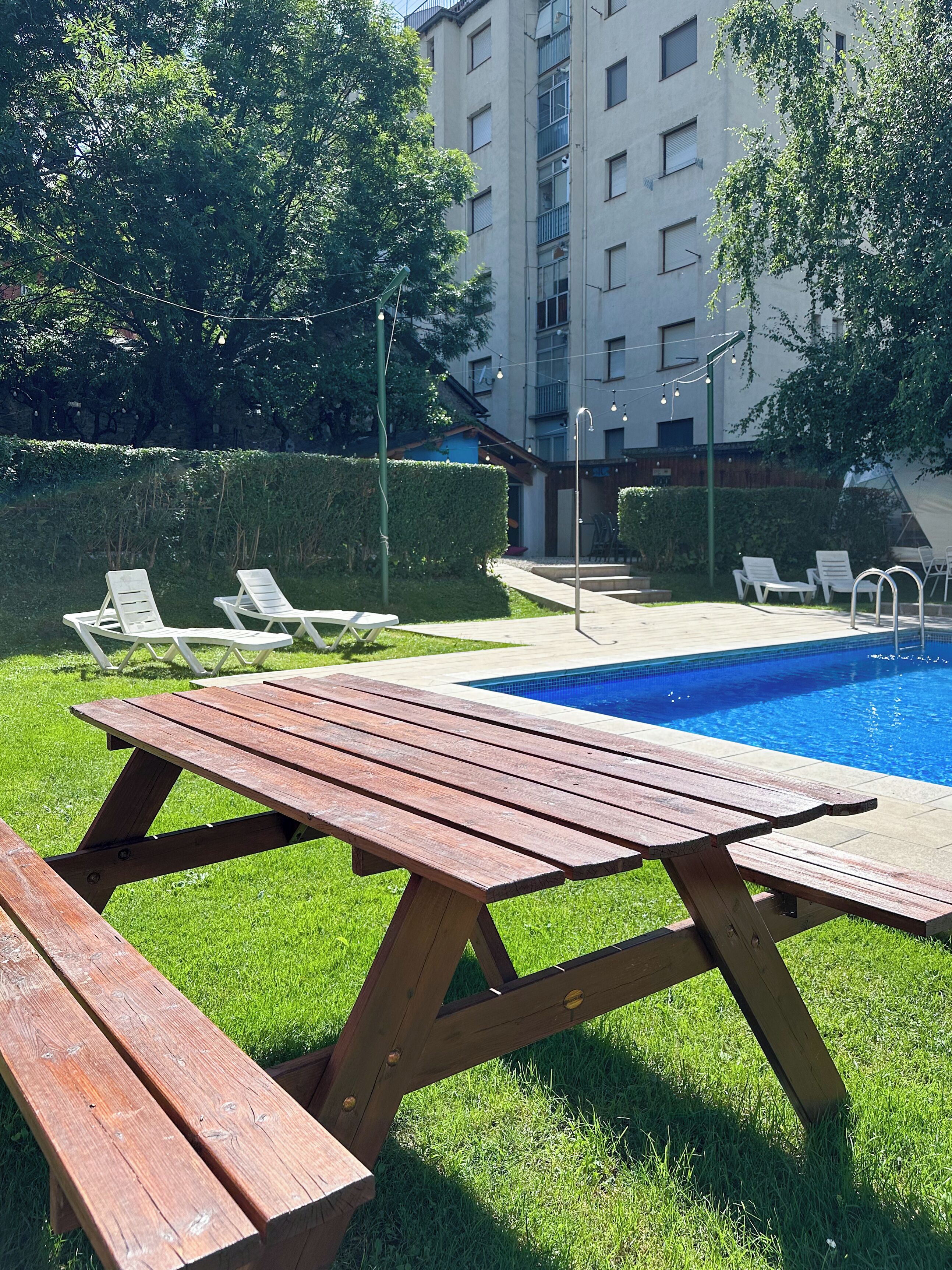 Zona de piscina con una mesa de madera y sillones de descanso, rodeada de un césped cuidado y un seto.
