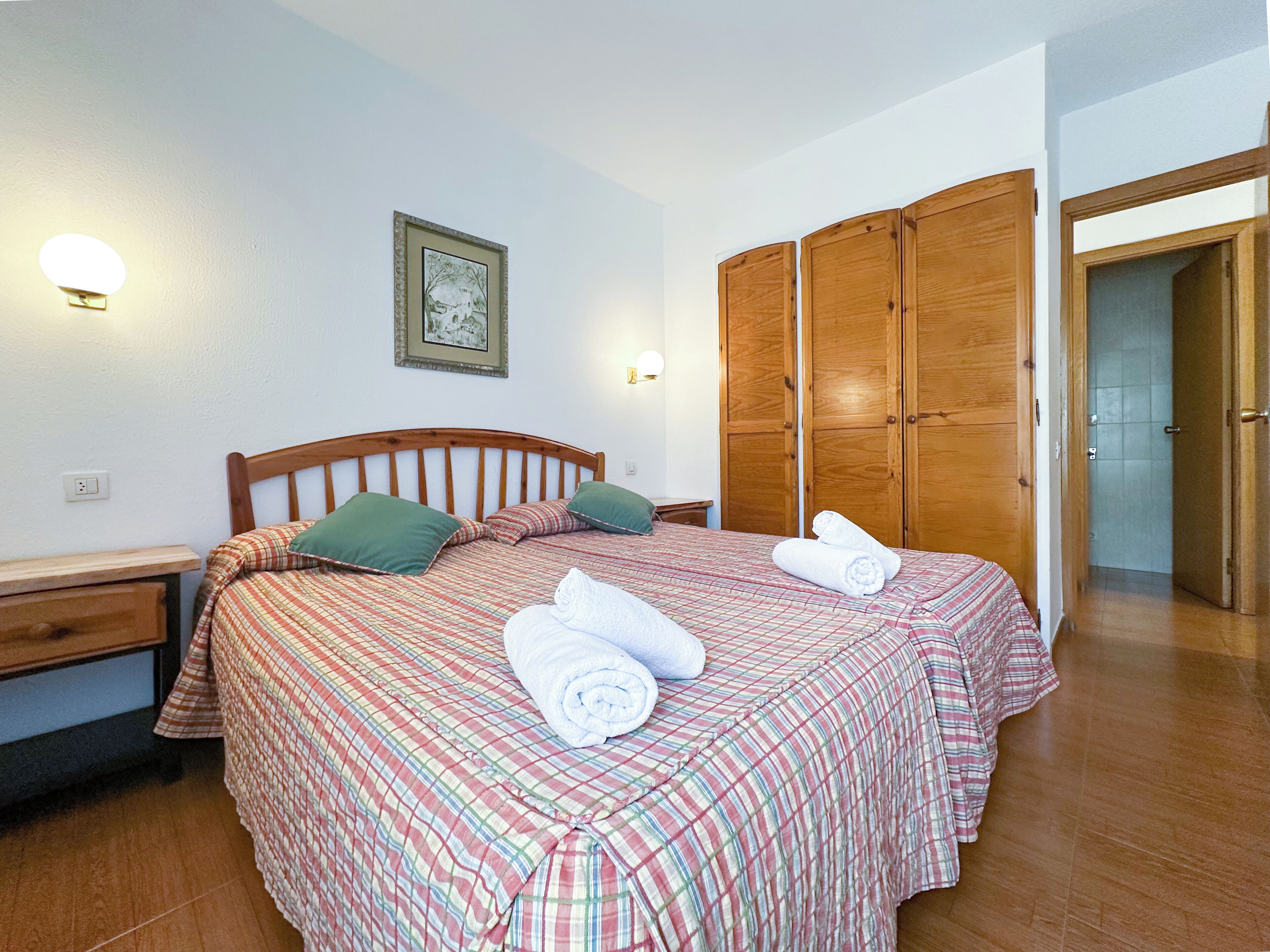 Un dormitorio con cama, armario de madera, mesita de noche y lámpara fijada a la pared.